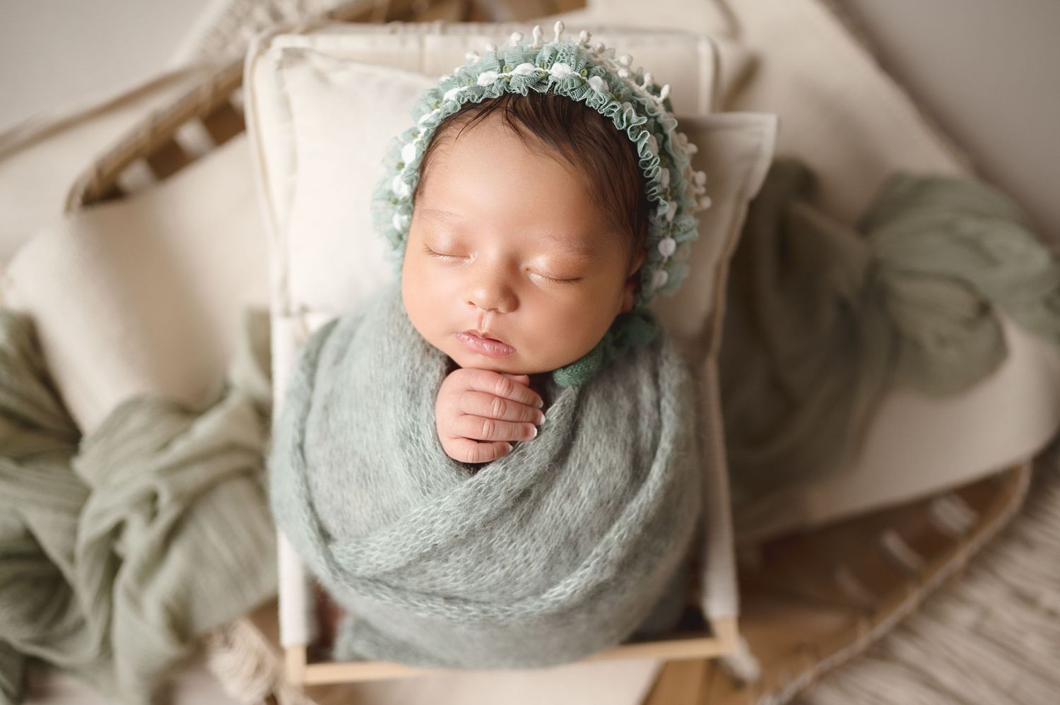 Newborn wrapped in soft mint green swaddle with floral crown against neutral background.