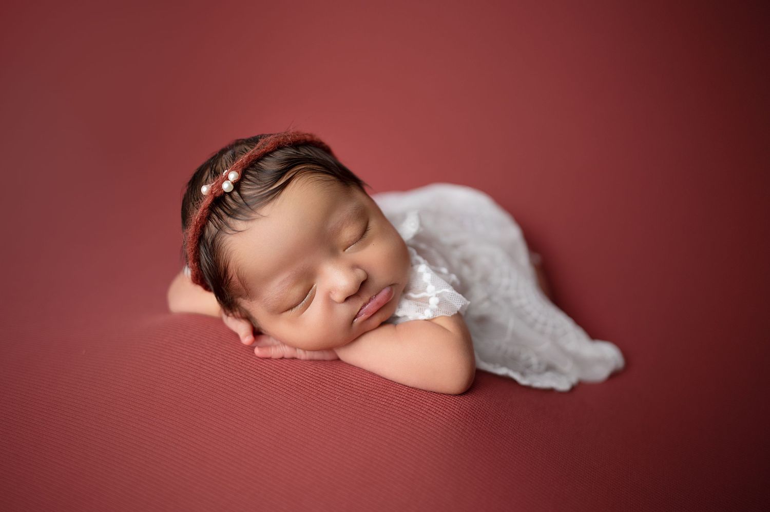 Sleeping newborn baby wrapped in white lace posed on a red background with delicate headband.