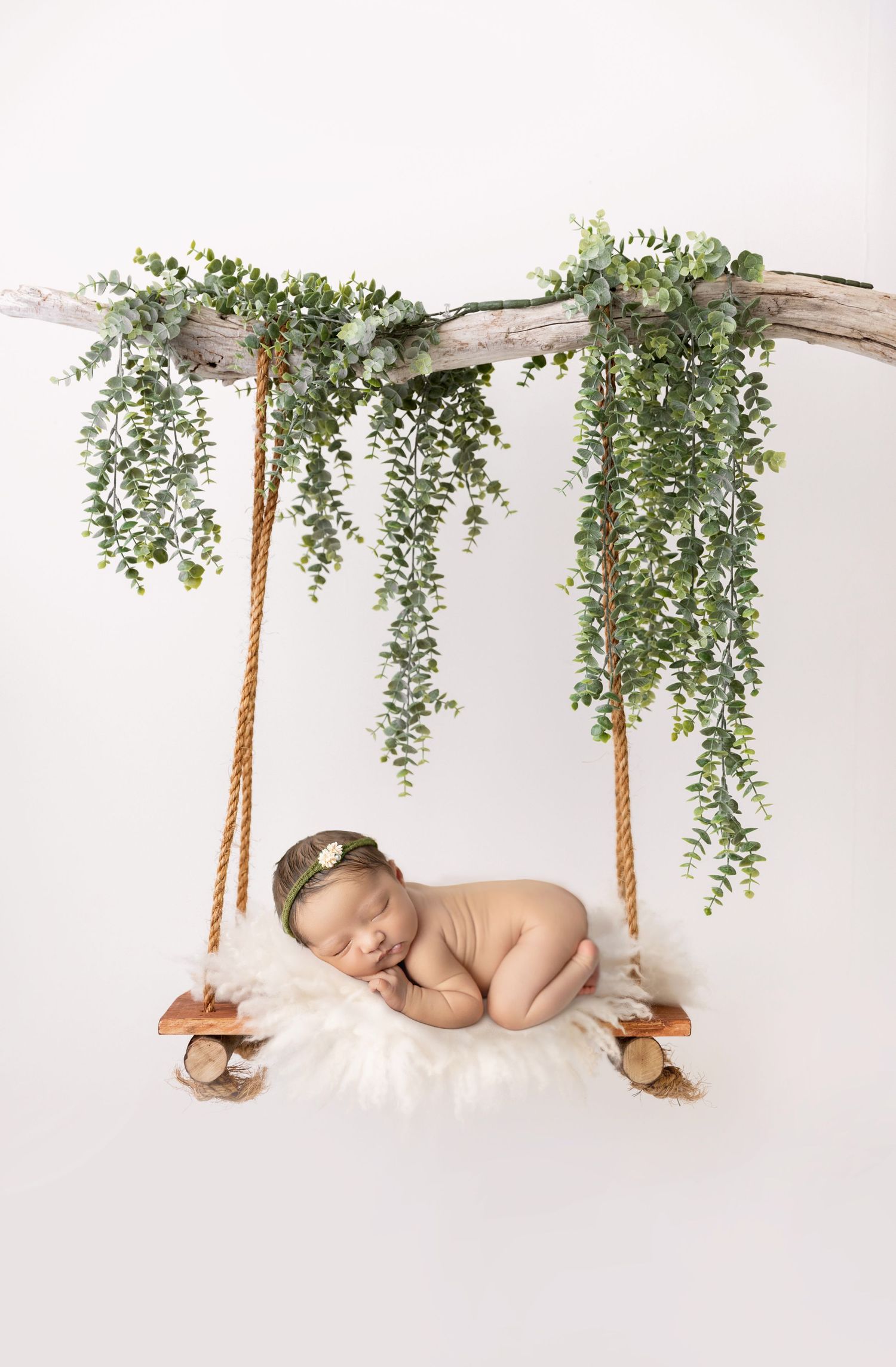 Sleeping newborn posed on a rustic wooden swing decorated with hanging greenery against white backdrop.