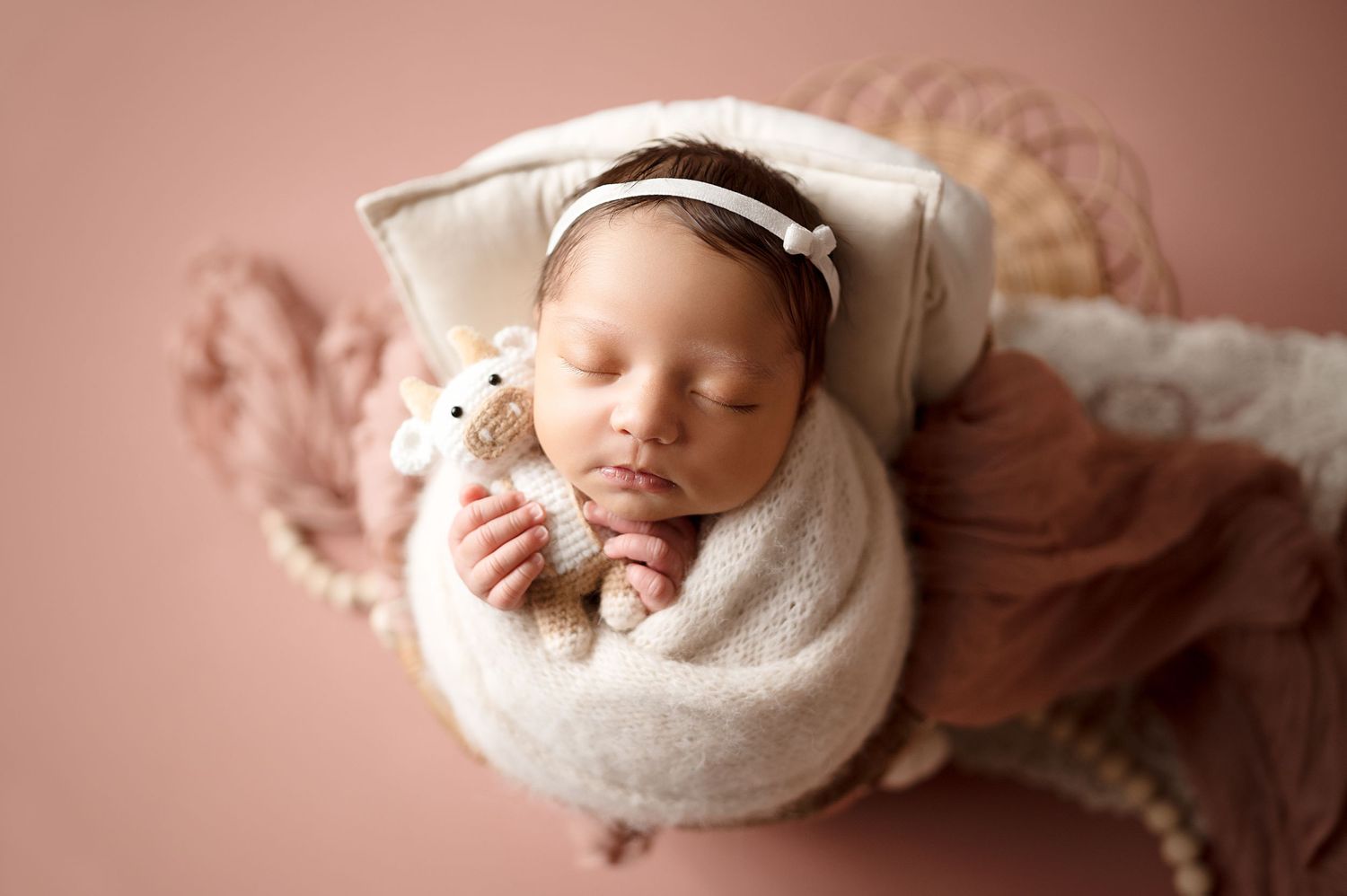Newborn baby in cream wrap sleeping peacefully in vintage styled setting with pink tones.
