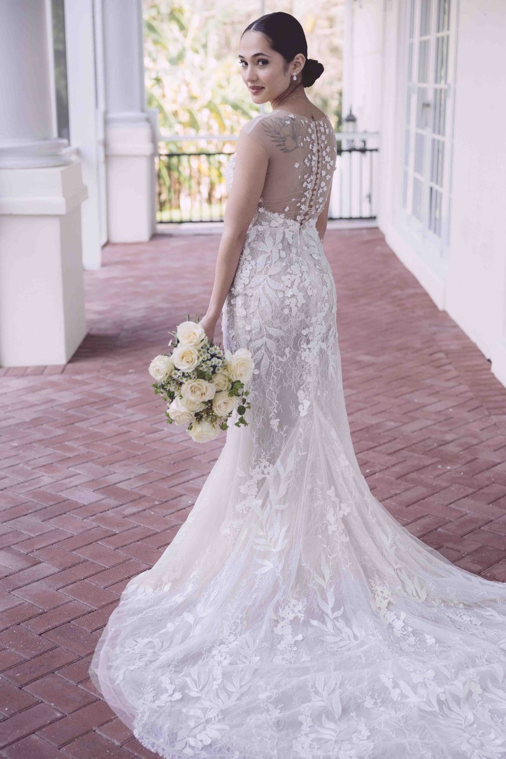An elegant lace wedding dress with illusion back detail and long train on brick patio.
