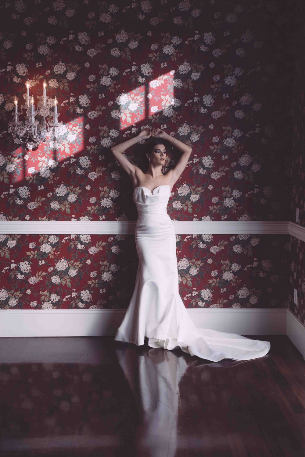 Elegant white wedding gown shown against dramatic red floral wallpaper with candlelit sconces.