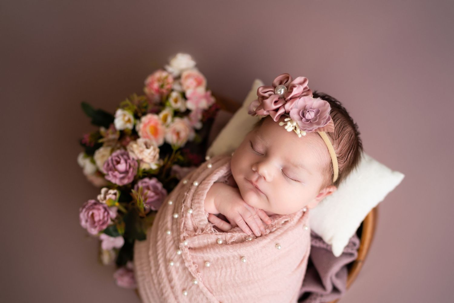 Sleeping newborn wrapped in pink lace with floral decorations on mauve background.
