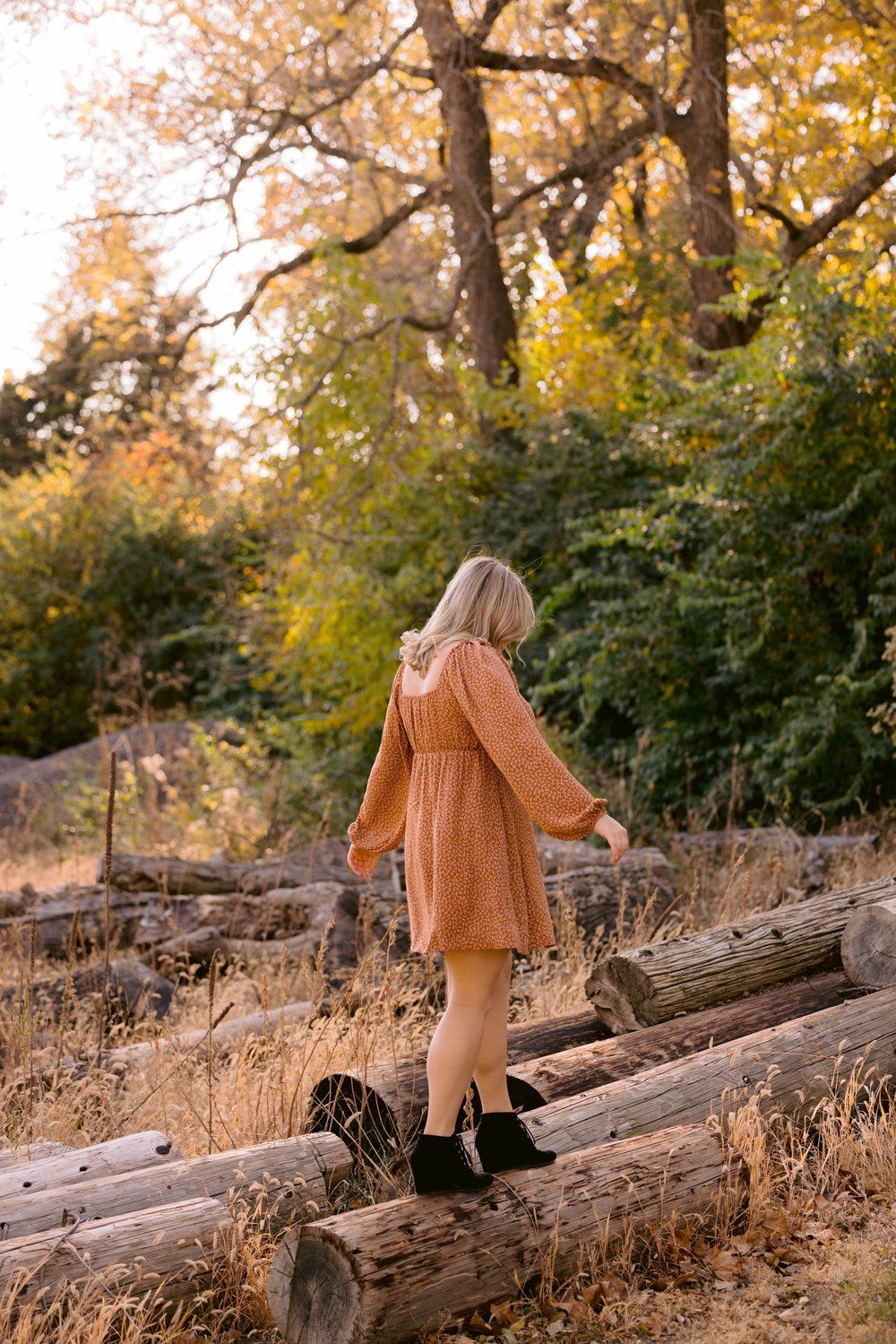 Person in orange sweater dress balancing on fallen log surrounded by autumn foliage.