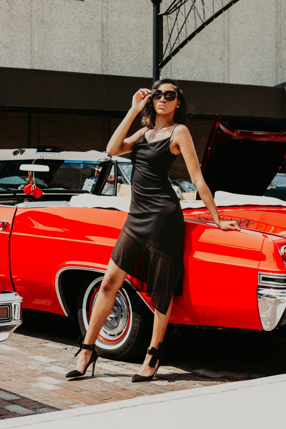 Stylish woman in black dress and sunglasses lounging against classic red automobile.