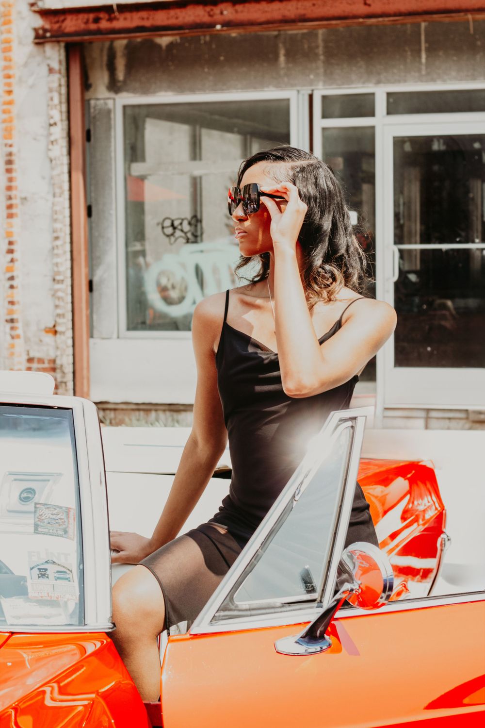 Woman in black dress posing next to vintage red car in urban setting.