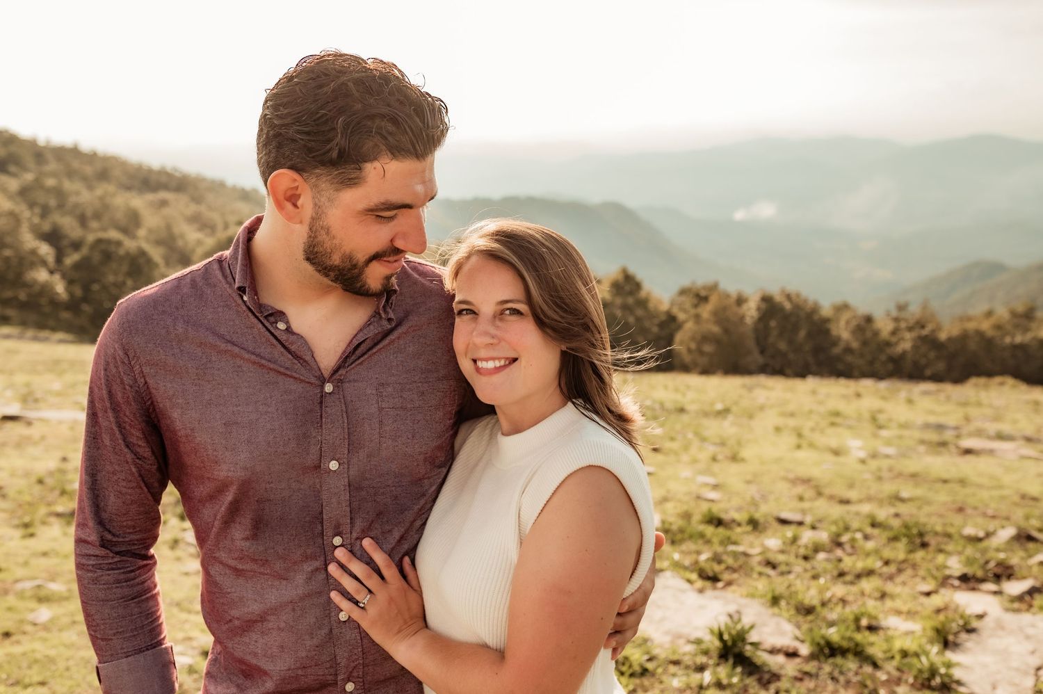 Woman looks at camera while man looks at her with Asheville mountains in background.