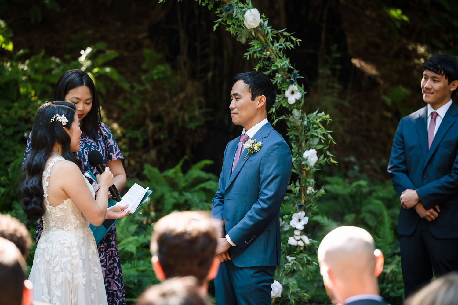 An outdoor wedding ceremony takes place beneath towering trees with floral arch decor.