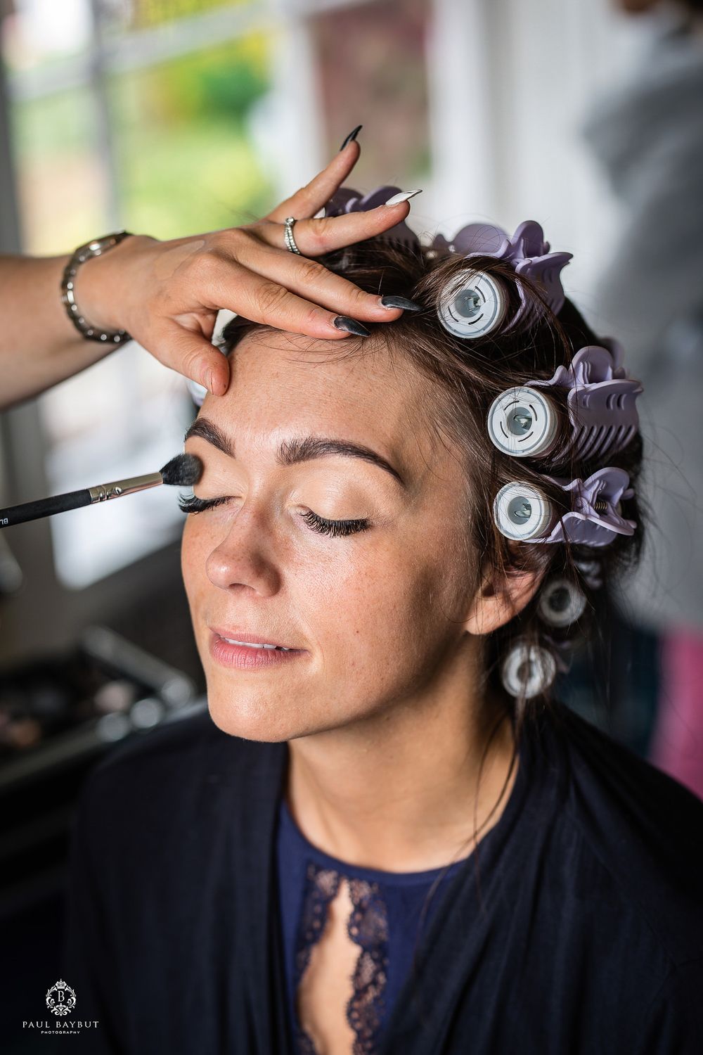 Makeup artist applies cosmetics to someone wearing hair rollers during wedding preparation.