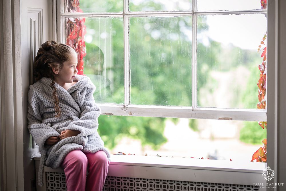 Child in gray sweater looking thoughtfully out window while sitting on windowsill.