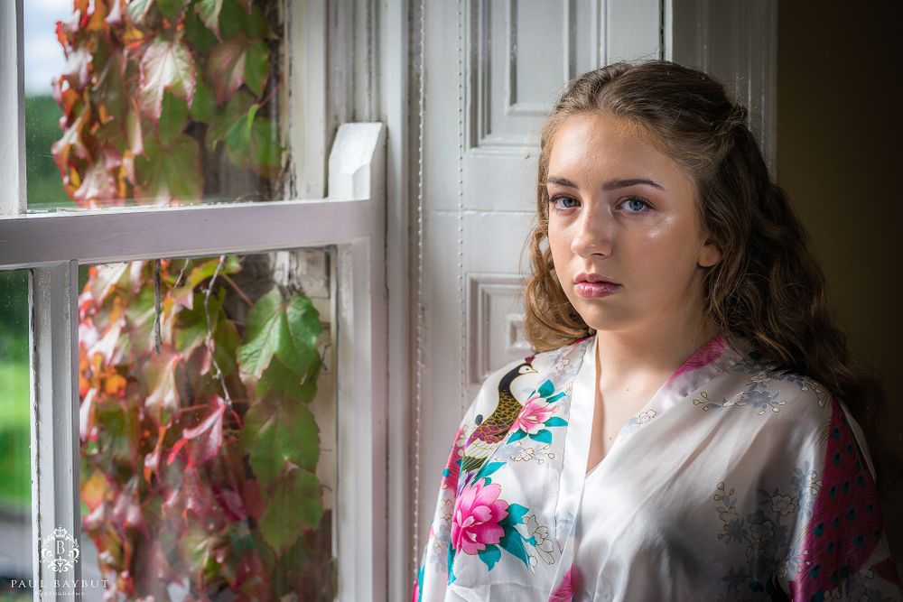 Person wearing white floral robe poses by window with fall foliage outside.