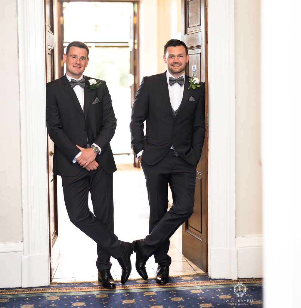 Two groomsmen in matching tuxedos pose casually in a doorway.