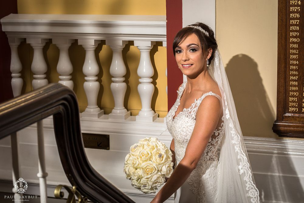 Bride in lace wedding dress holds white bouquet while posing near ornate banister.