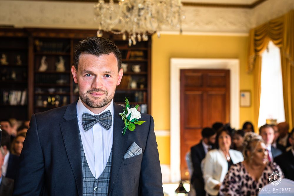 Groom in navy tuxedo and bow tie stands at altar during wedding ceremony.