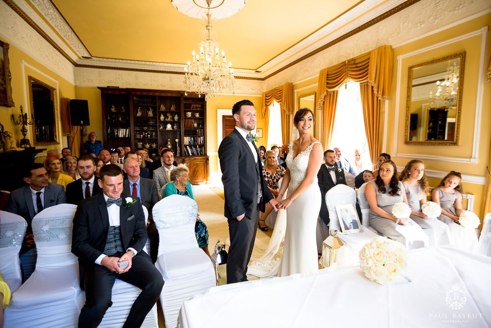 Wedding ceremony in a bright yellow room with chandelier and white-covered chairs.