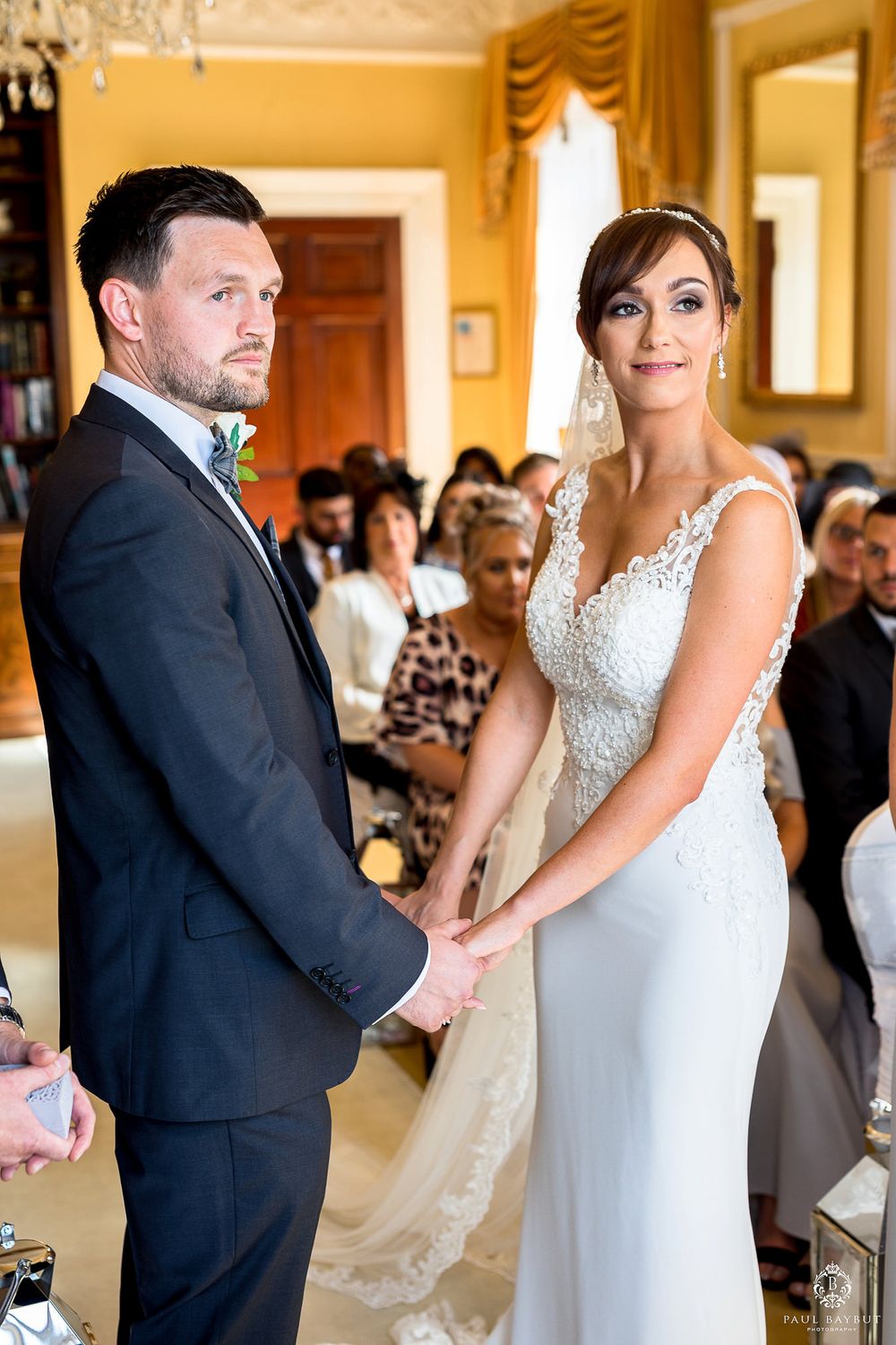 Couple holding hands during wedding ceremony in elegant yellow room with chandelier.