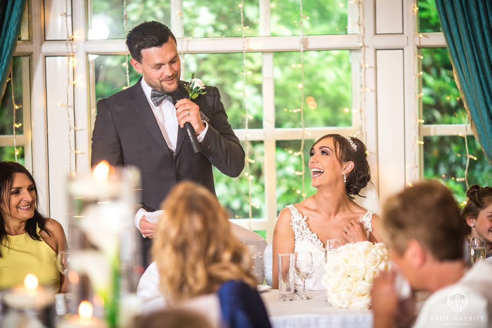 Wedding reception speech moment with guests seated at decorated tables by large windows.