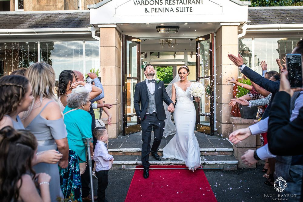 Newlyweds exit their wedding venue on a red carpet as guests throw confetti in celebration.