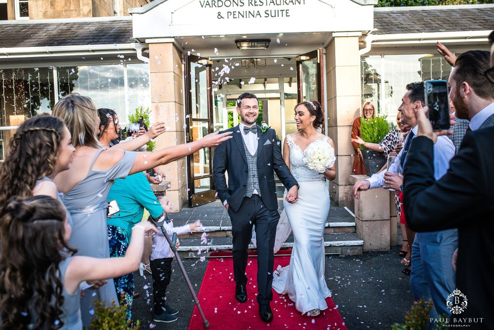 Wedding guests toss confetti at a couple walking down a red carpet outside a venue entrance.