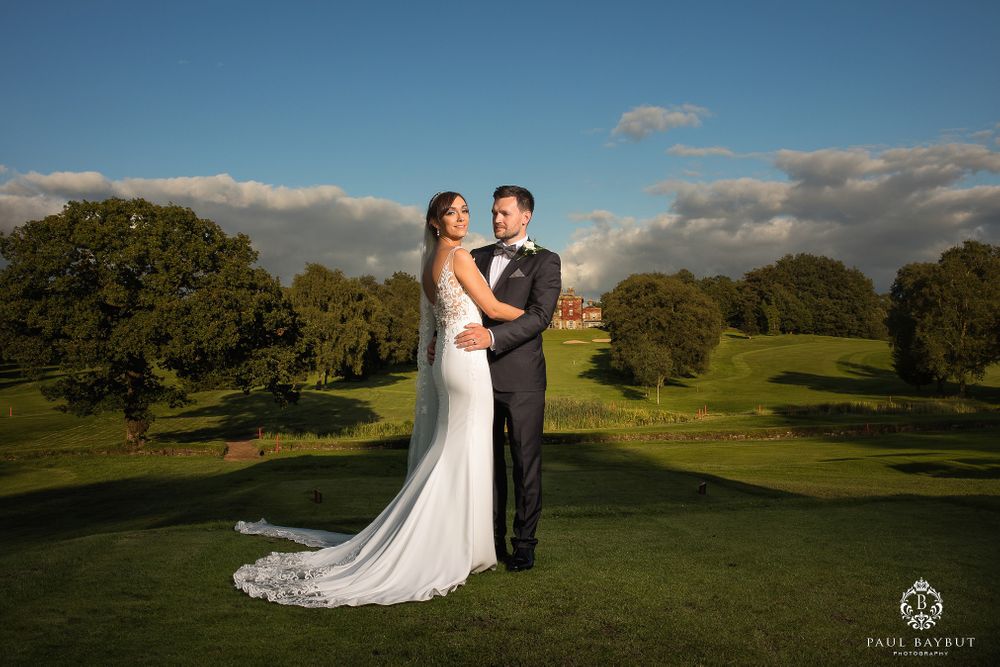 Elegant wedding couple poses on a lush green golf course with trees in the background.