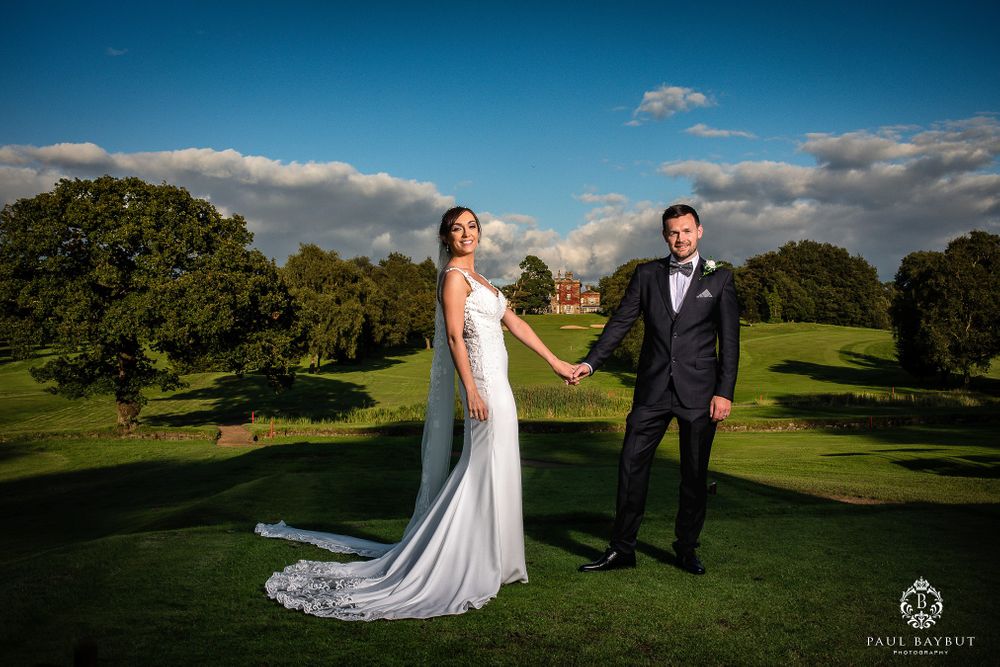 Bride in fitted gown and groom in tuxedo pose on manicured golf course grounds.