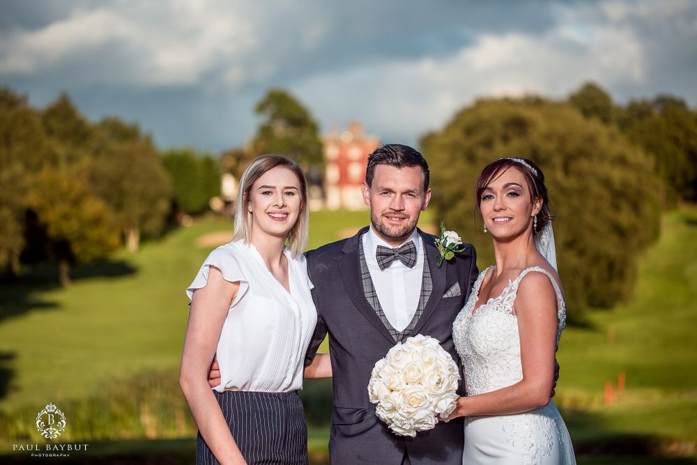Wedding party poses on manicured grounds with historic building in background.