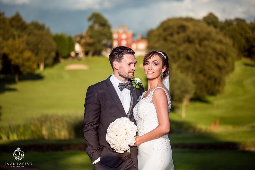 Elegant wedding couple poses on lush green grounds with a stunning manor house visible in the background.