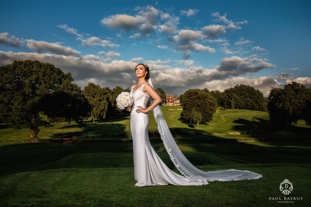Bride in flowing white gown holds bouquet while standing on manicured lawn under dramatic sky.