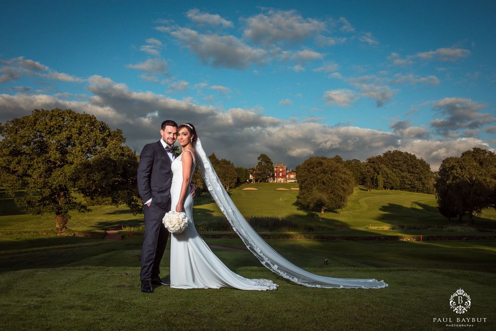 Newlyweds embrace on grassy hillside with dramatic long veil flowing in evening light.