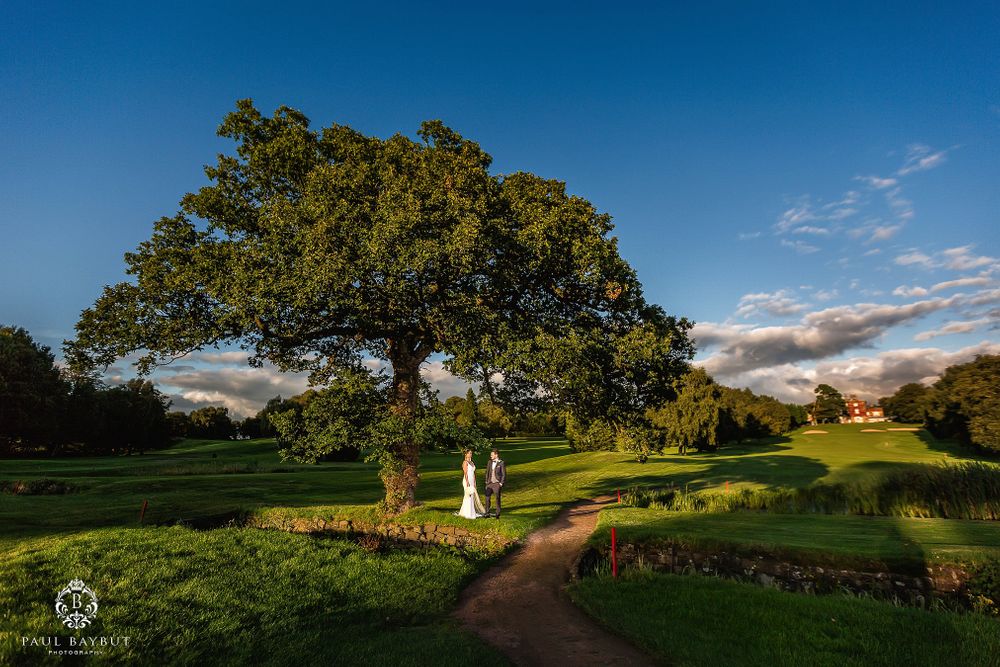 Wedding couple walks distant garden path under large oak tree at sunset.