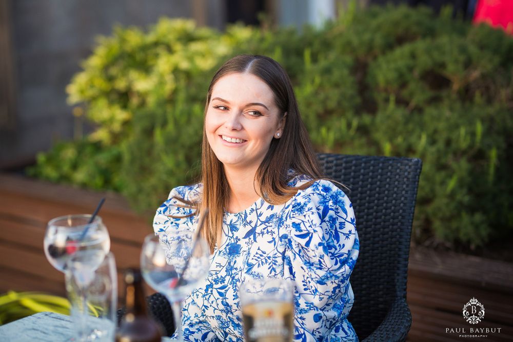 Woman in blue and white patterned dress smiles while seated at outdoor dining table.