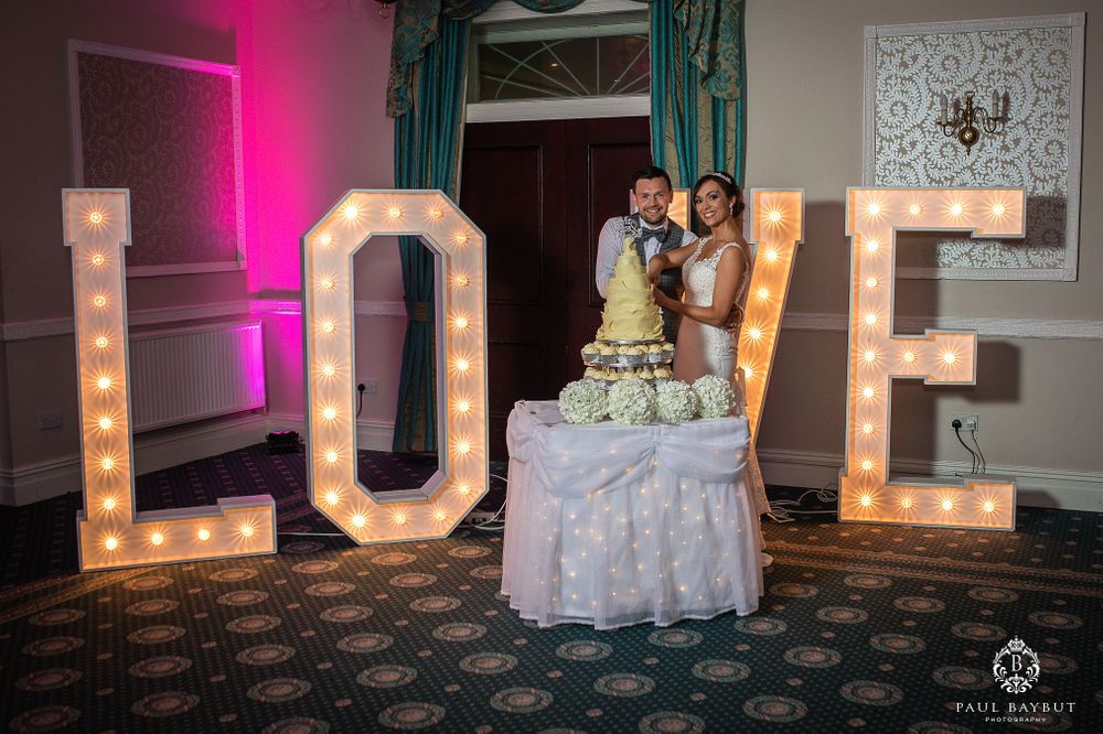 Wedding cake displayed in front of illuminated LOVE marquee letters with teal curtains in background.