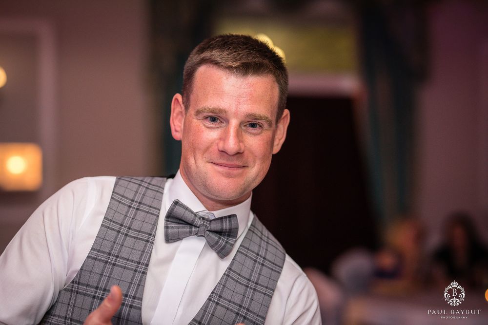 Well-dressed person in grey vest and bow tie smiling at wedding reception.