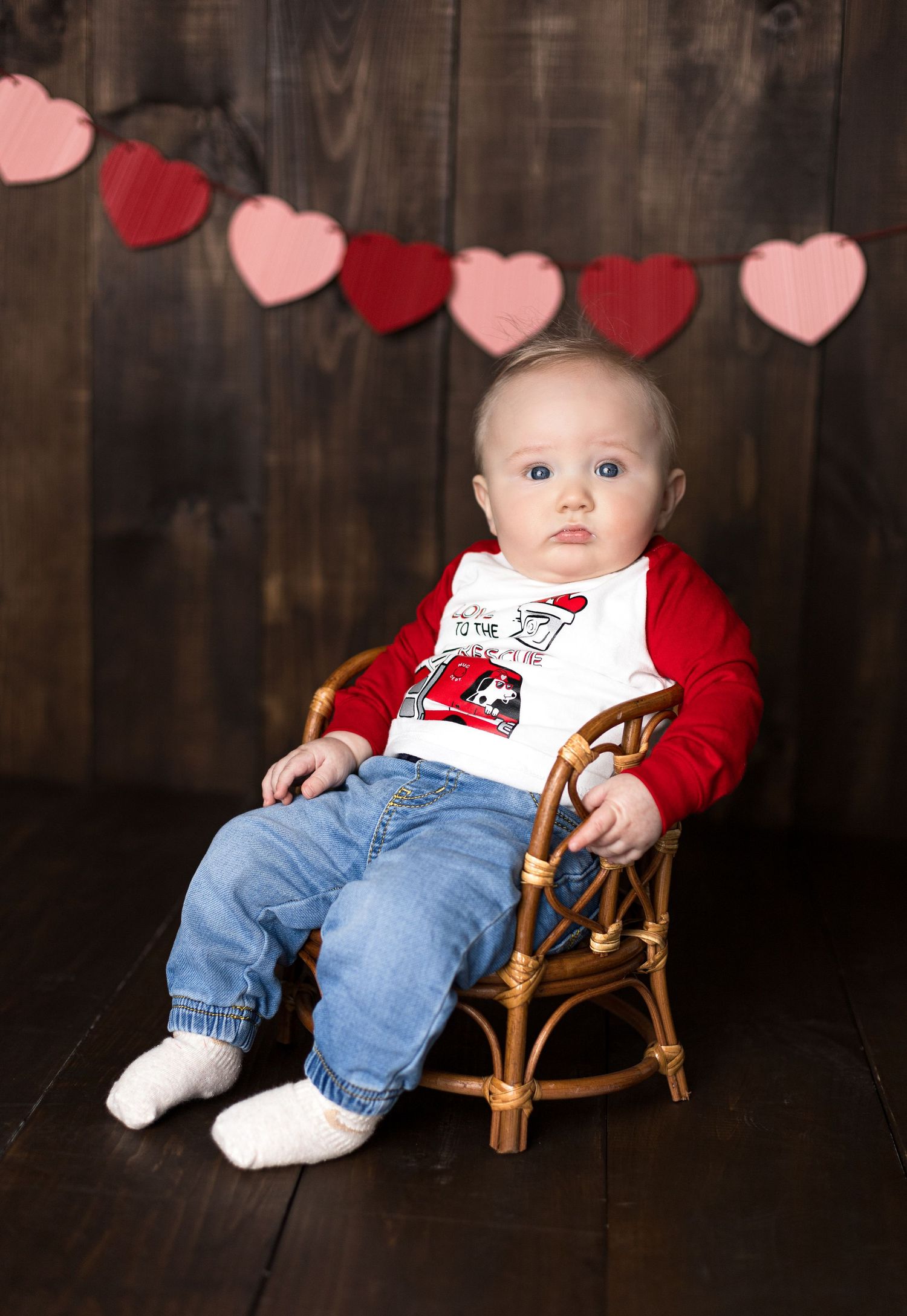 Baby sitting in wooden chair wearing red and white outfit with heart banner decoration.