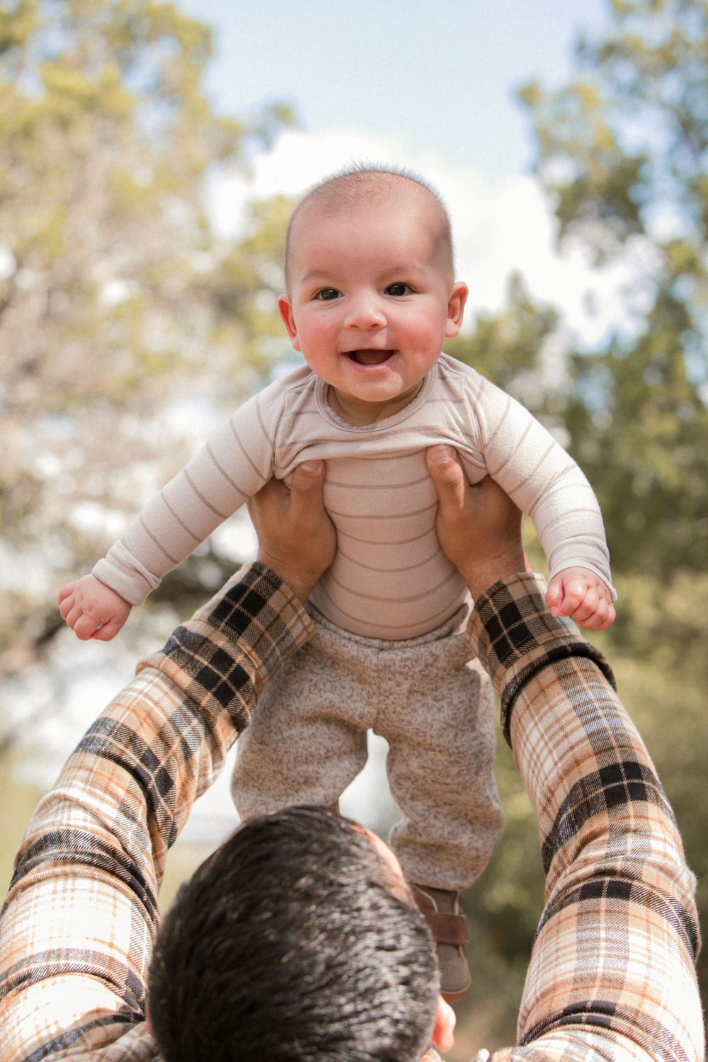Baby wearing cream colored outfit being lifted playfully in the air against a sunny background.