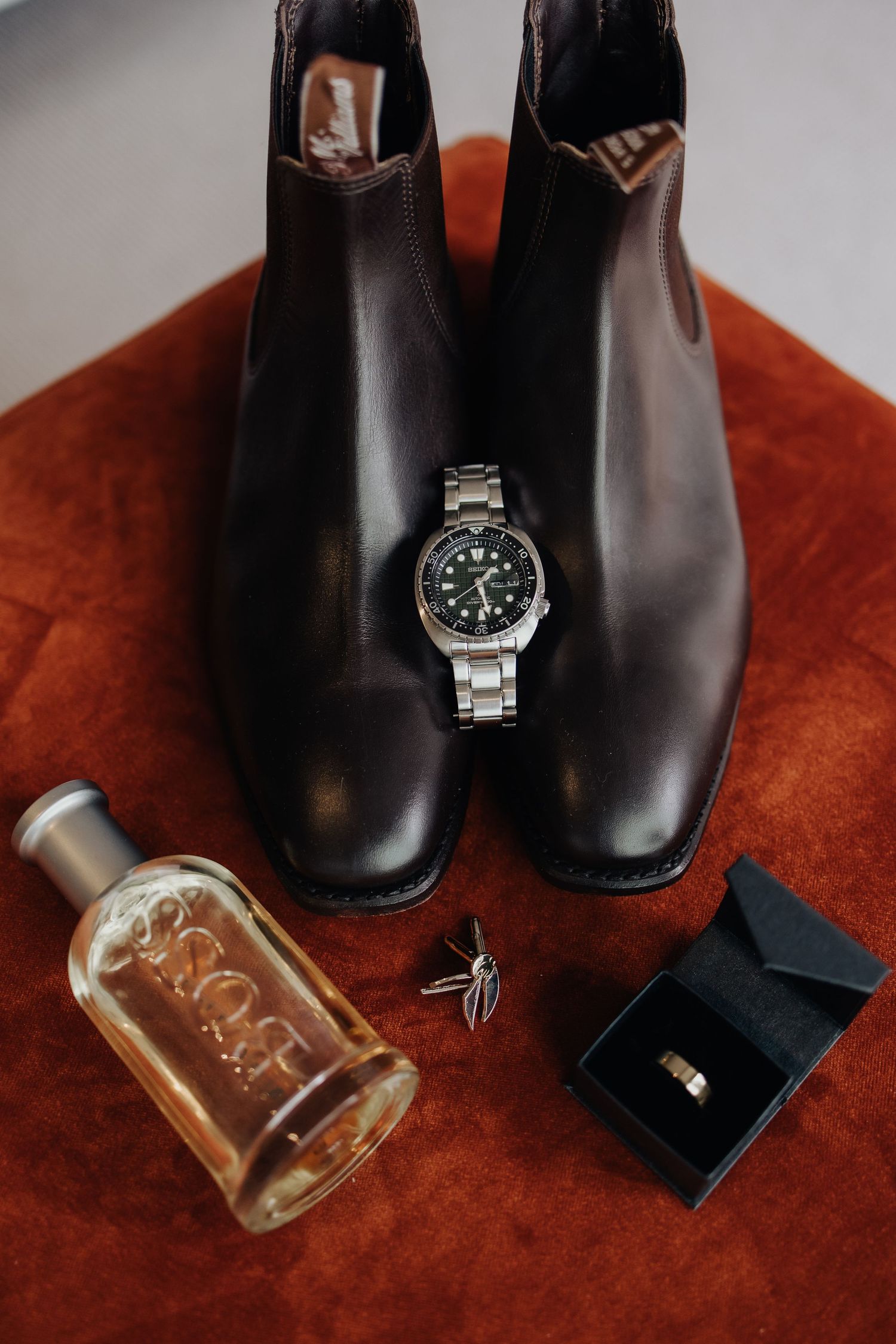 Wedding accessories including black boots, watch, and perfume bottle arranged on red velvet surface.