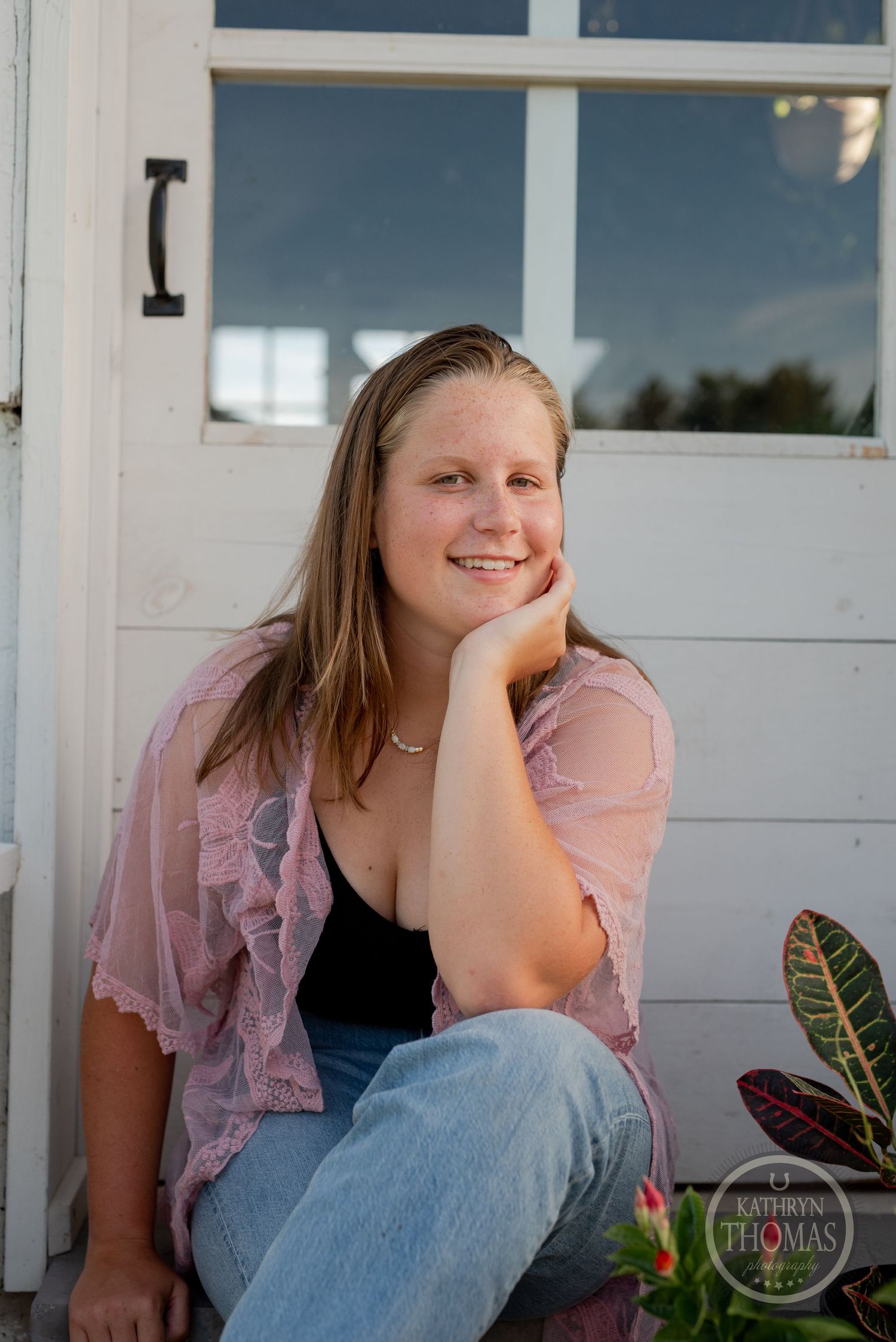 Portrait of someone leaning against a white door wearing a pink lace top and jeans in outdoor setting.