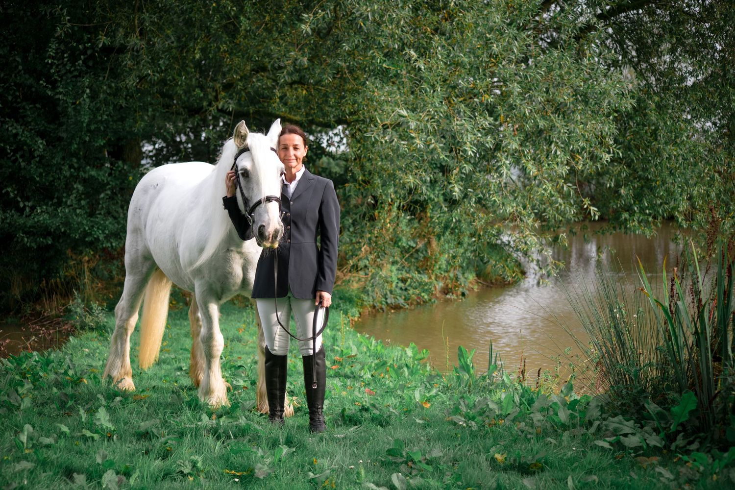 A white horse and rider stand beside a tranquil pond in a lush green setting.