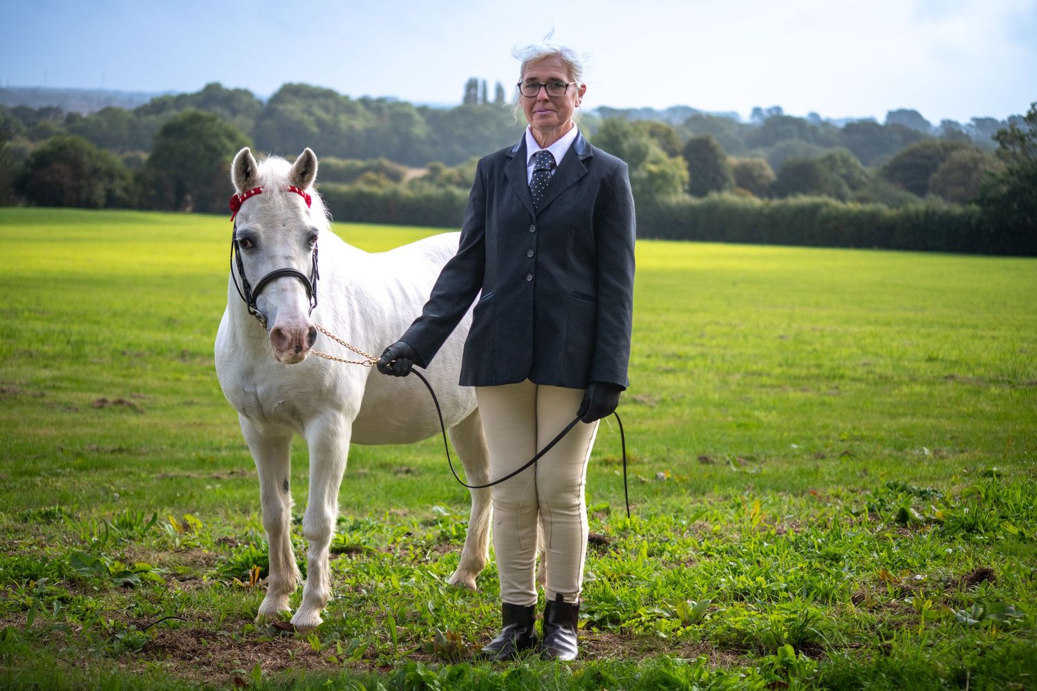 White pony and rider stand in green pasture with rolling hills in background.