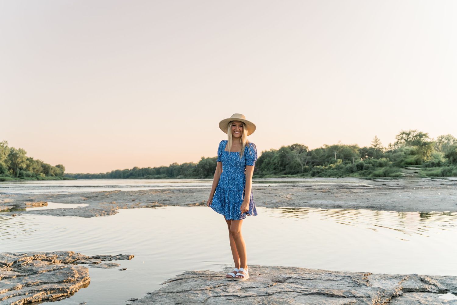 Natural light senior portrait of teen in vibrant blue dress and tan hat on the river’s edge in Maumee.