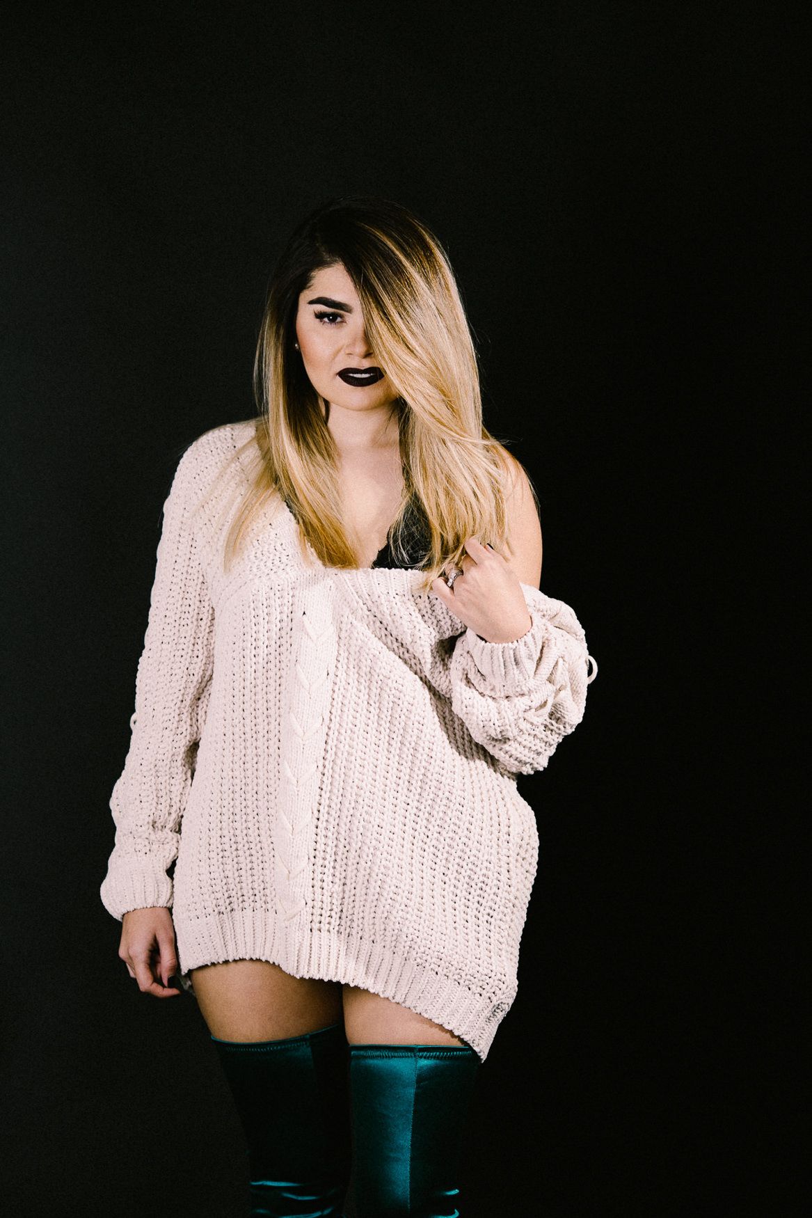 Fashion model in oversized beige sweater and dark boots poses against black background.