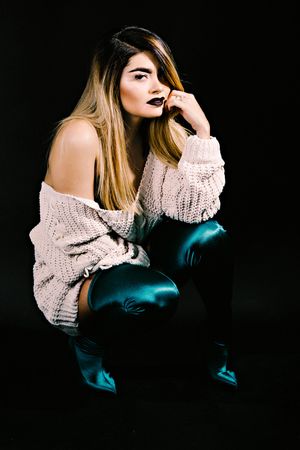 Creative fashion portrait with model in cream sweater and turquoise boots against dark backdrop.