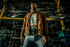 Person wearing brown leather jacket and jeans standing in industrial setting with mechanical equipment.