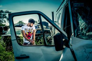 Person in graphic t-shirt sitting in driver's seat of vehicle against blue sky background.