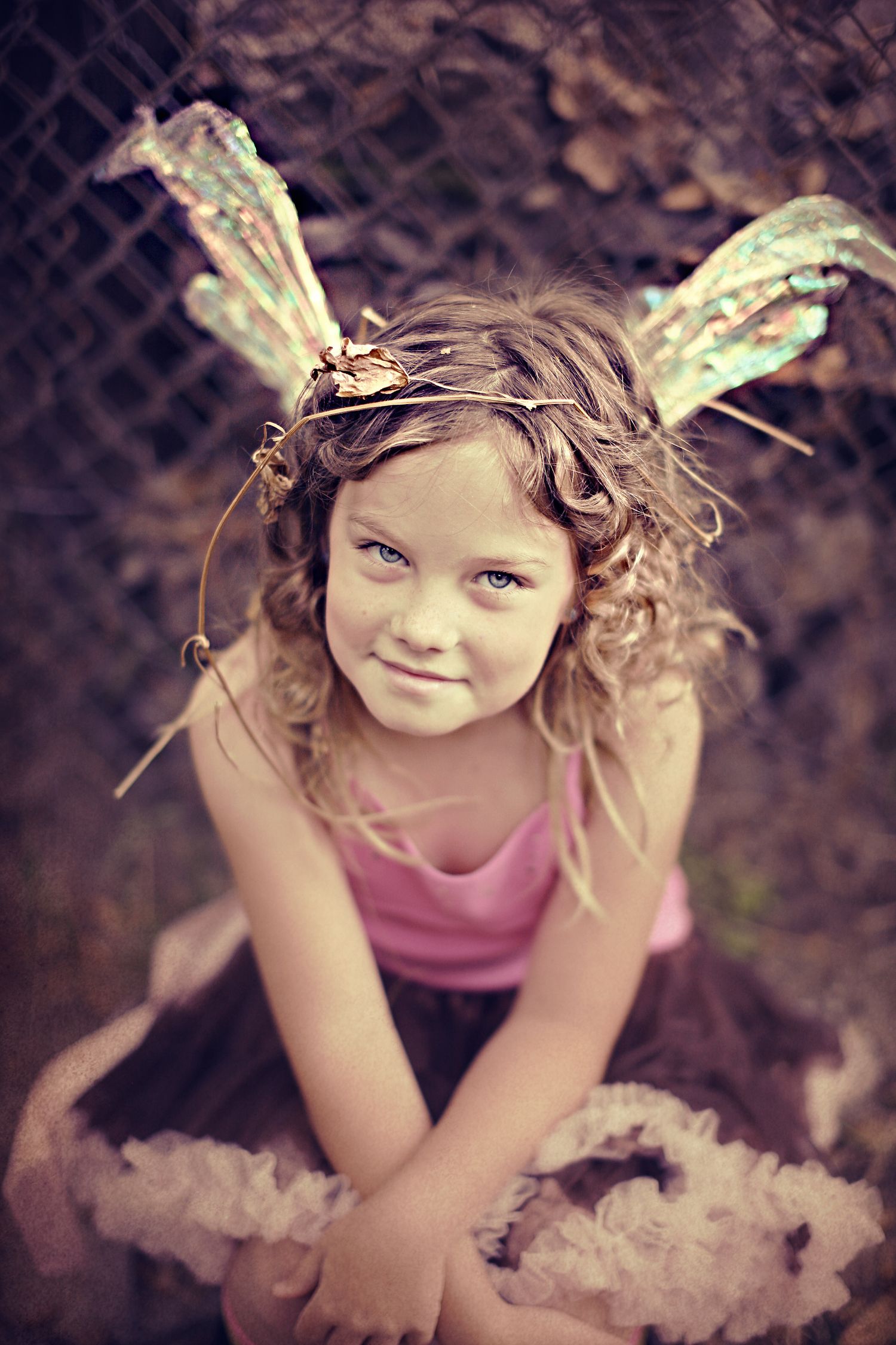 A young child in a pink top and tutu wears sparkly fairy wings and poses outside near a dark fence.