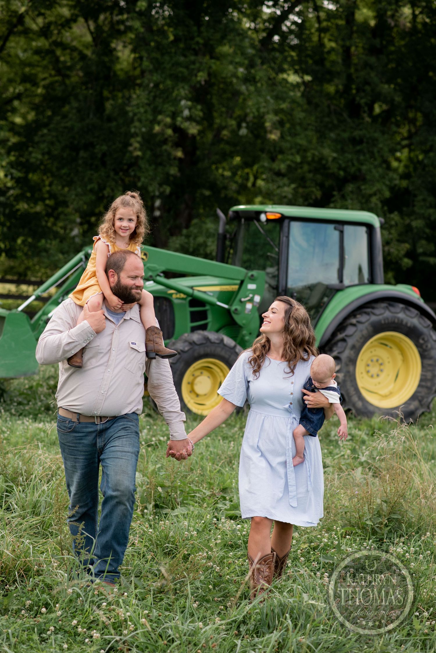 A happy family poses together in front of their green John Deere tractor in a grassy field.