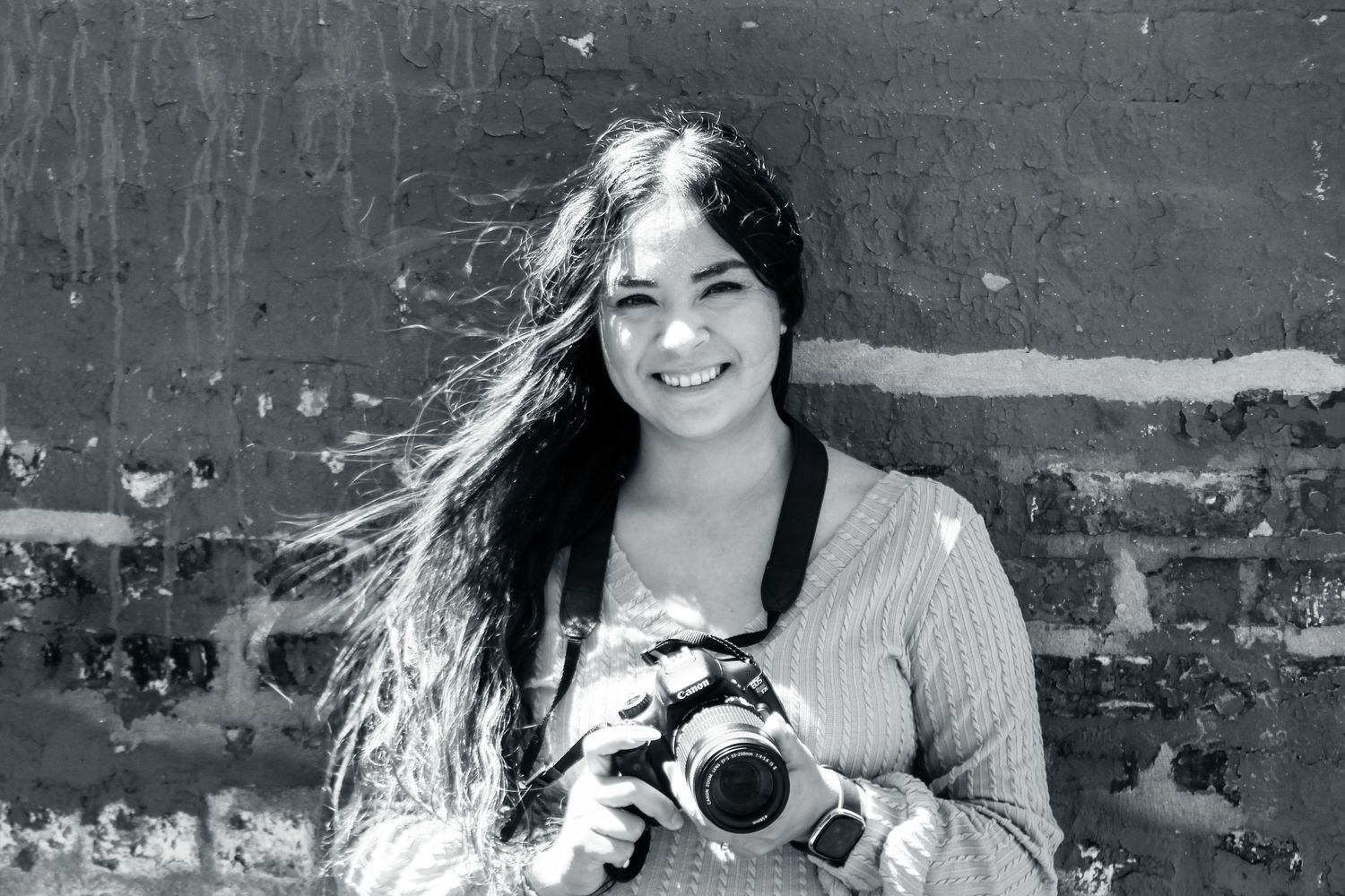 A photographer with long dark hair smiles while holding a vintage camera against a textured wall.