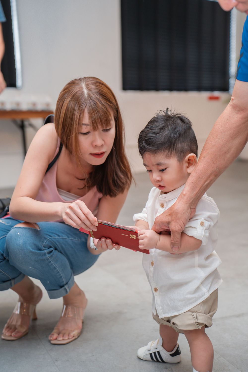 Adults help a small child in white clothing learn to use a red item in an indoor setting.