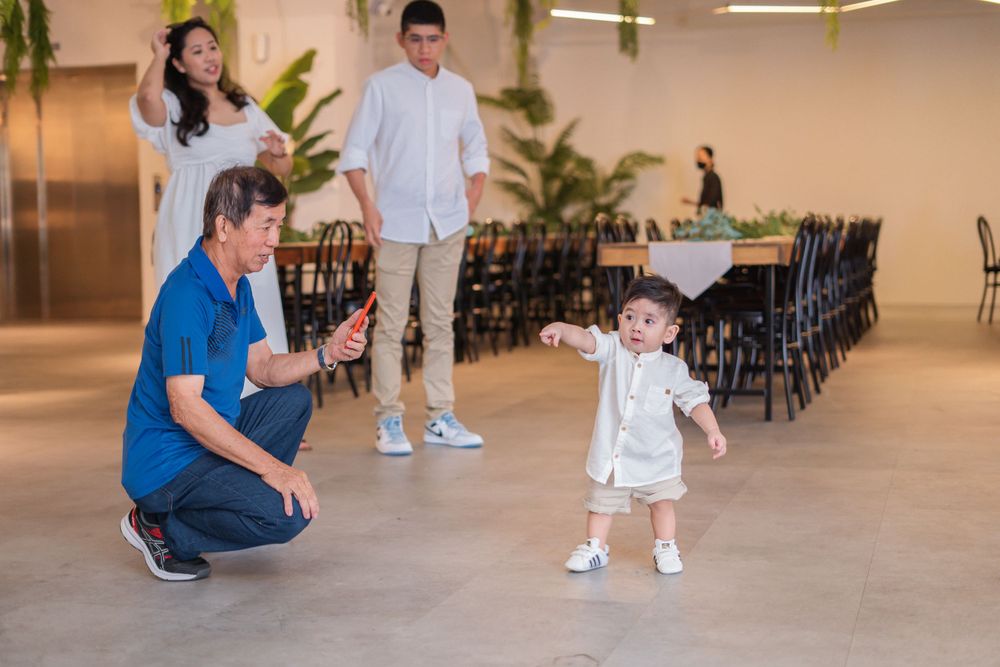 Young toddler takes first steps while adults watch encouragingly in a spacious indoor venue.