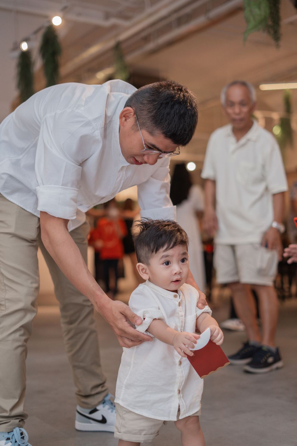 Adult guides small child holding red packet in white outfit at indoor event.